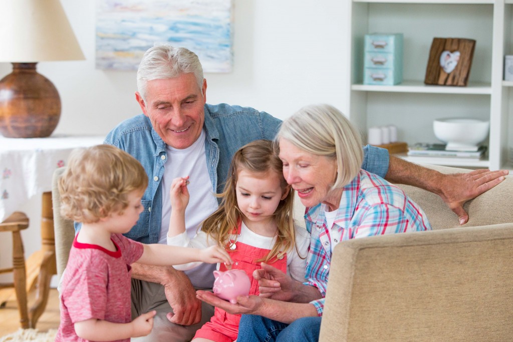 Two happy young children put some money into a piggy bank with their grandparents. They are sitting on the sofa in their home together wearing casual clothing.